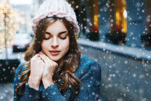 winter-haut-rau-cosmetics-trockene-haut-problemhaut Frau mit warmer Mütze, die in winterlicher Umgebung steht und die Hände vor der Brust hält, während Schnee fällt, symbolisiert die Notwendigkeit von Hautpflege im Winter.