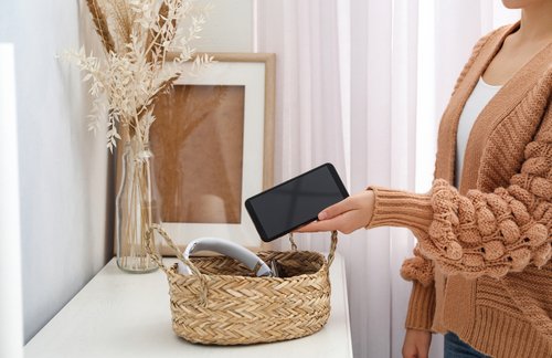 Person legt Smartphone in einen geflochtenen Korb und betont damit digitale Entgiftung und reduzierte Gerätenutzung, mit einer ruhigen häuslichen Umgebung und dekorativen Elementen.