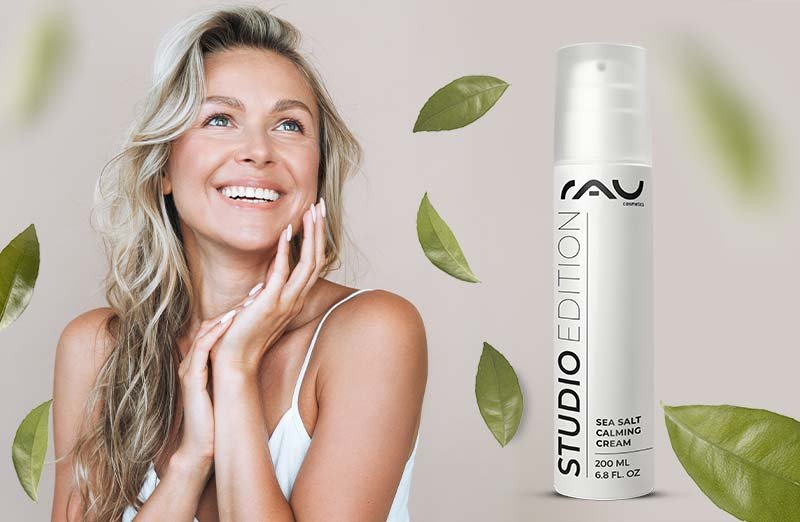 Lächelnde Frau mit Studio Edition Sea Salt Calming Cream und grünen Blättern im Hintergrund für Hautpflege.