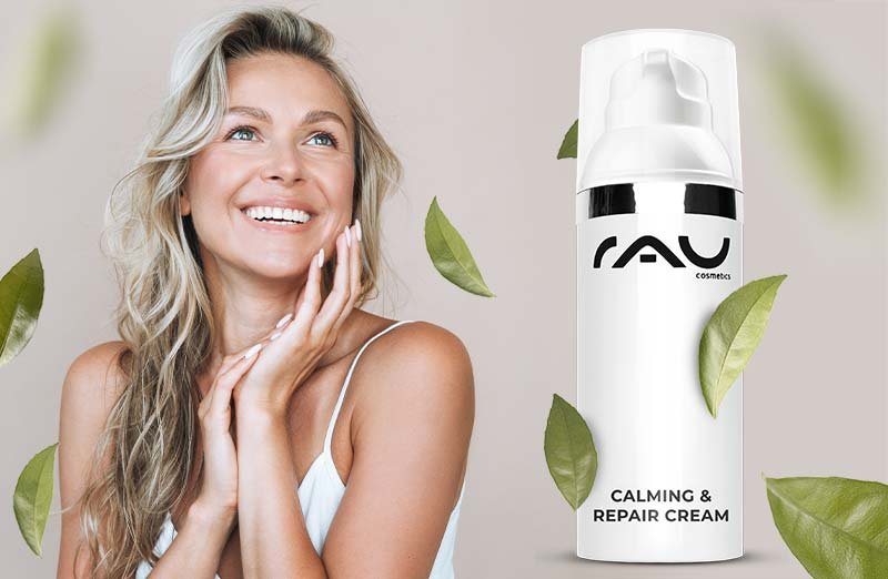 Blonde Frau trägt die RAU Cosmetics Calming & Repair Cream auf, im Hintergrund sind zarte grüne Blätter zu sehen.