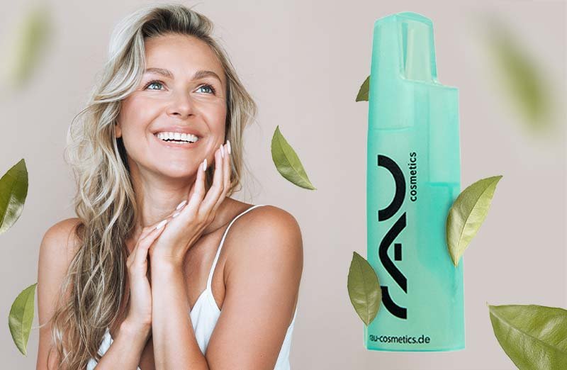 Lächelnde Frau mit langen, lockigen Haaren und grünem RAU Cosmetics Flasche, umgeben von grünen Blättern.
